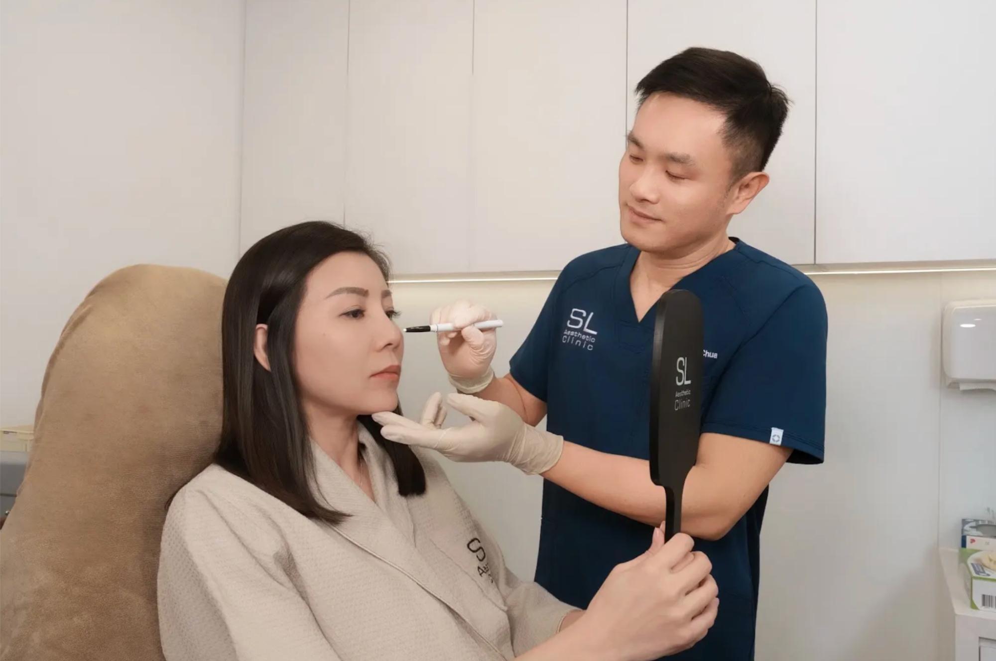 Aesthetic doctor performing a facial treatment on a seated woman while she looks into a handheld mirror