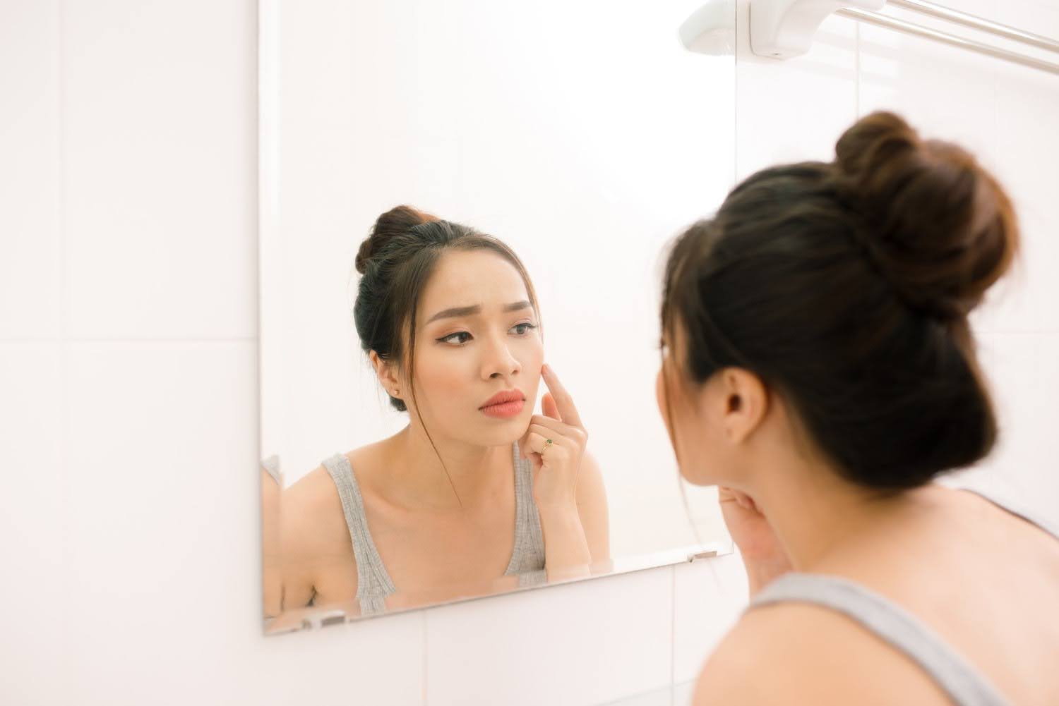 Woman examining her cheek in a bathroom mirror with a concerned expression